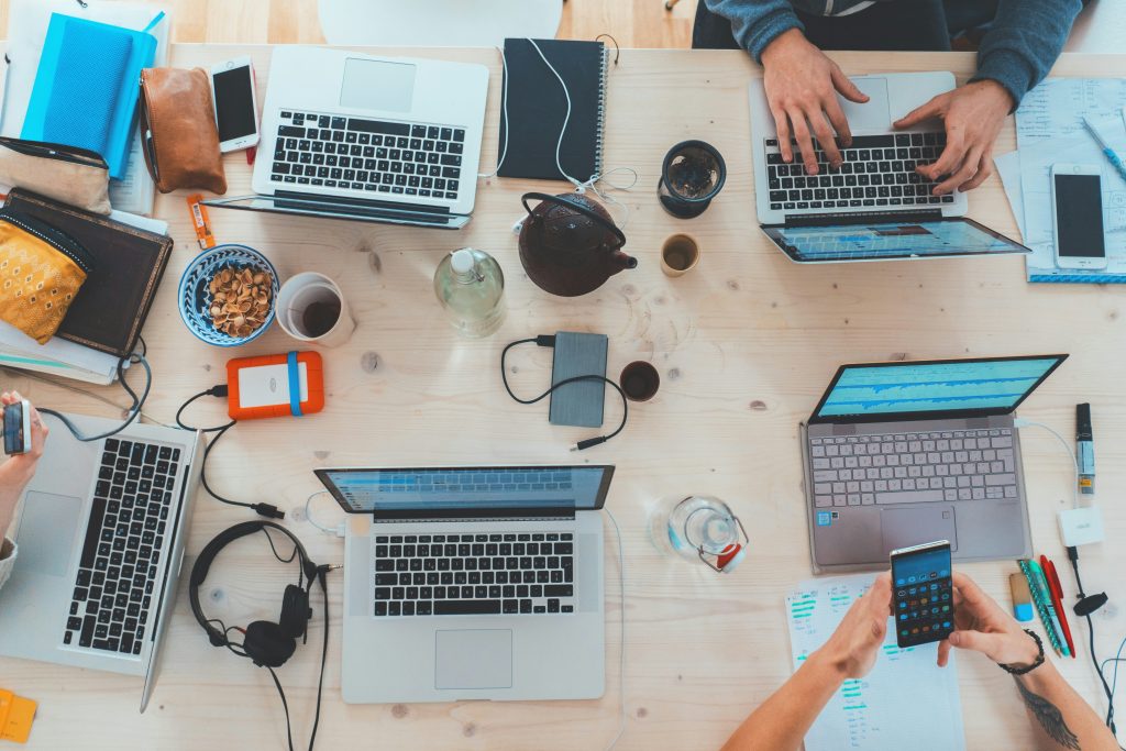 Men working on computers across a desk with cereal in a bowl and a Chinese cast iron teapot with coffees. One holds a mobile phone.